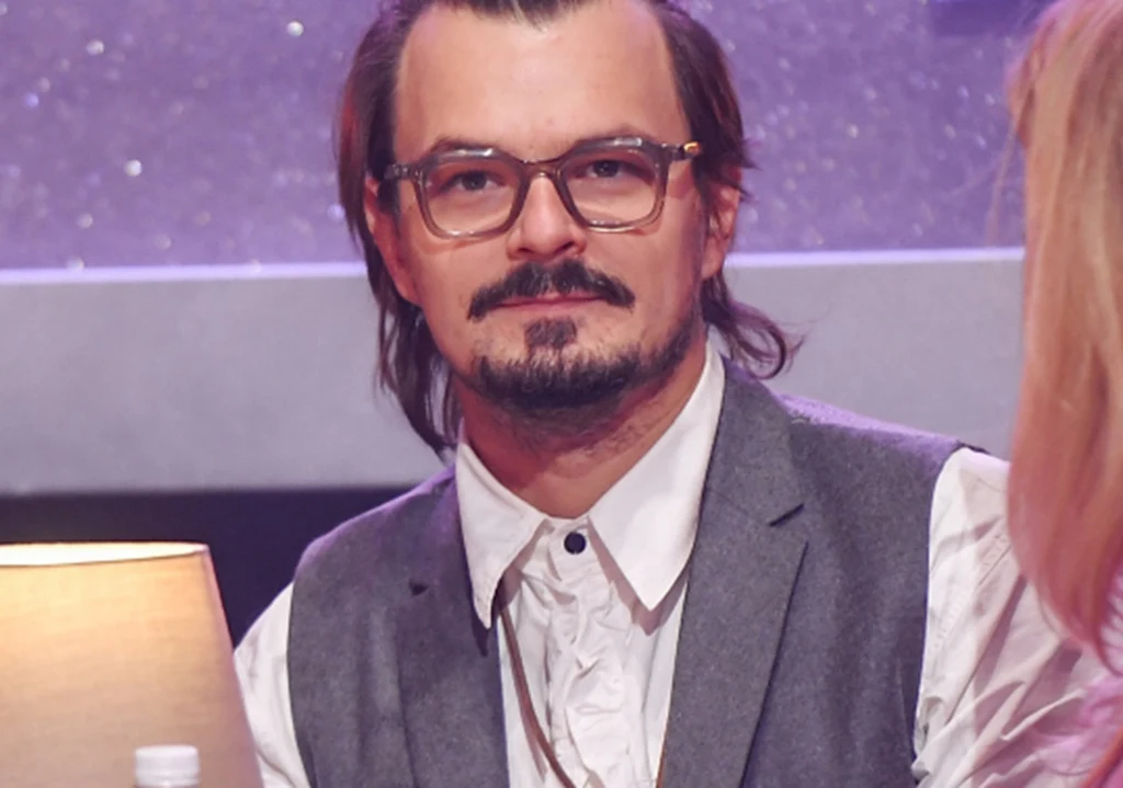 Dawid Ogrodnik Mężczyzna w okularach i szarej kamizelce siedzi przy stole, patrzy prosto w obiektyw, po prawej stronie widoczna jest kobieta odwrócona tyłem, na pierwszym planie stoi lampa.