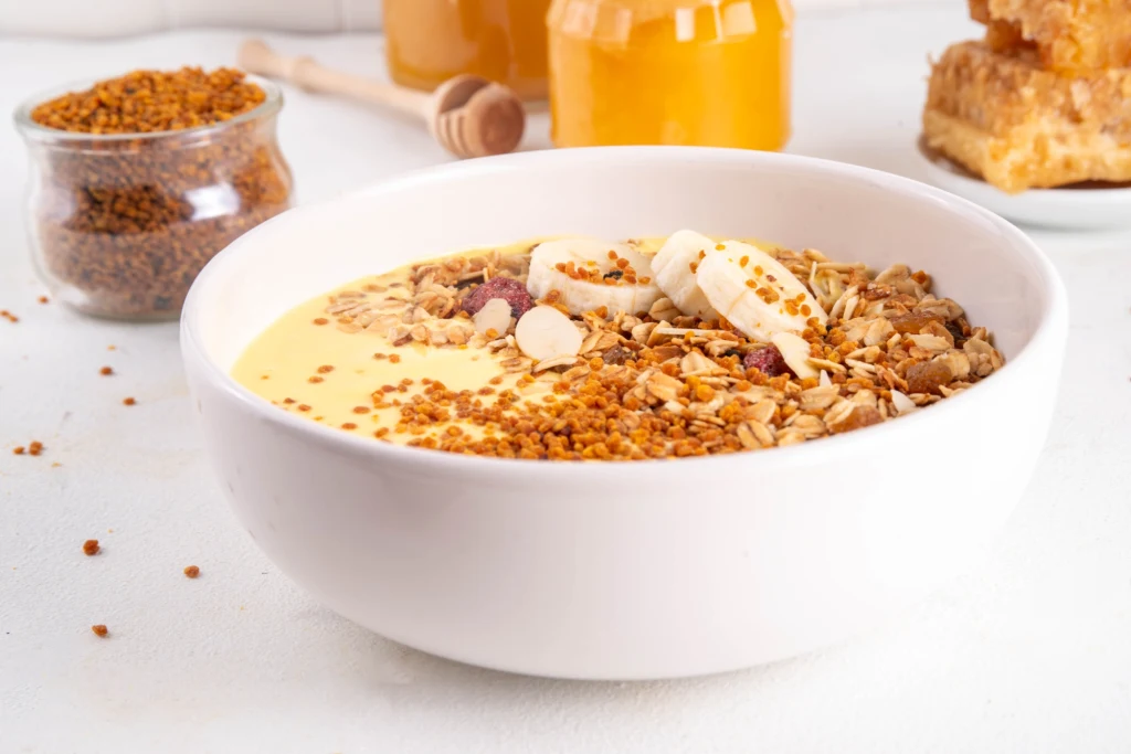 Czym jest pierzga i dlaczego warto ją spożywać? Biała miska wypełniona owsianką z musli, plasterkami banana oraz dodatkiem miodu i pyłku pszczelego. W tle słoik z miodem, kawałek plastra miodu oraz drewniana łyżeczka do miodu.