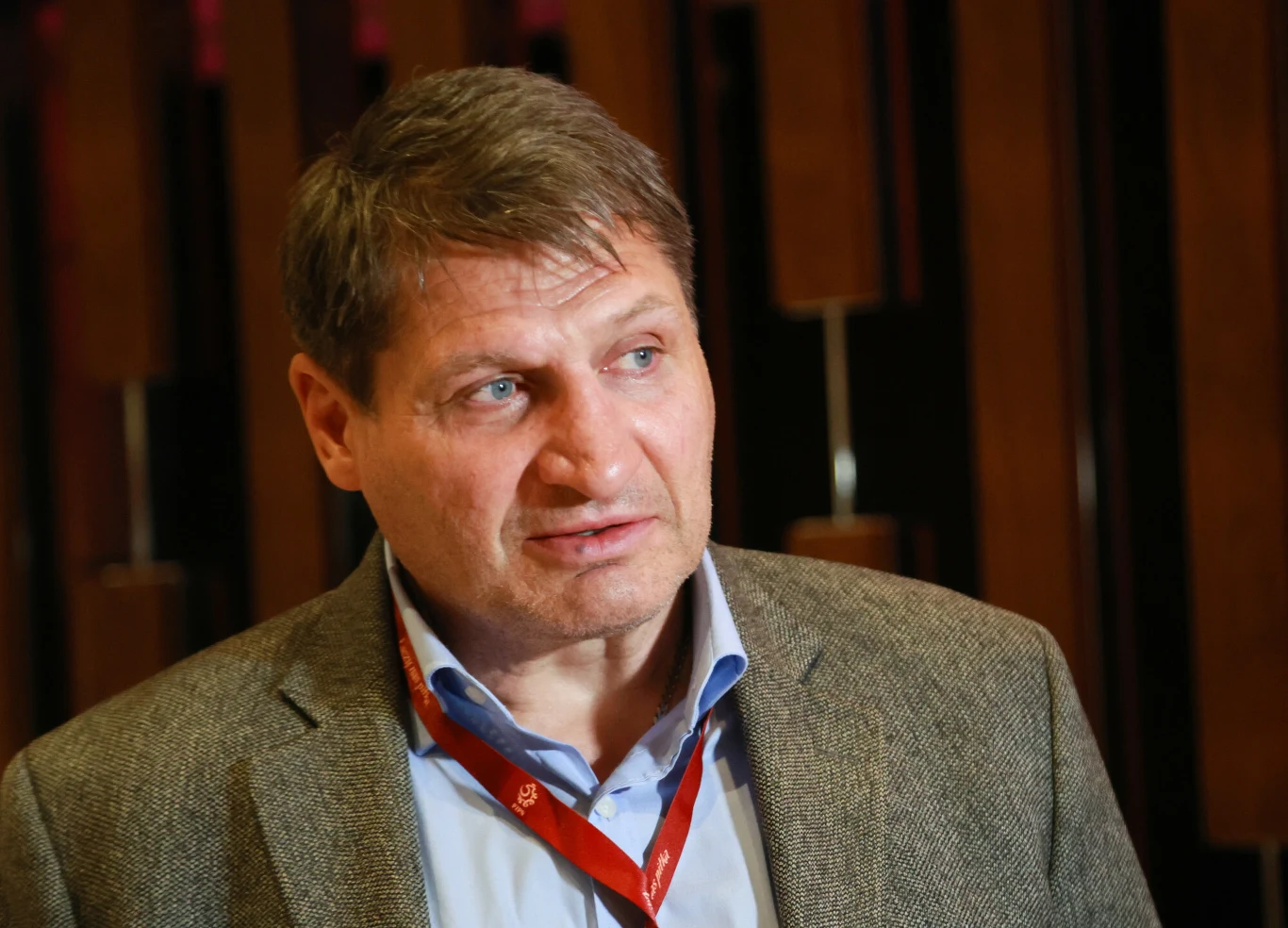 Mężczyzna w średnim wieku w marynarce i niebieskiej koszuli, z czerwonym identyfikatorem na szyi, na tle drewnianych paneli.