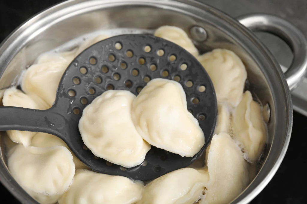 Pierogi w garnku podczas gotowania, podnoszone łyżką cedzakową. Dumplings unoszą się na powierzchni wody.