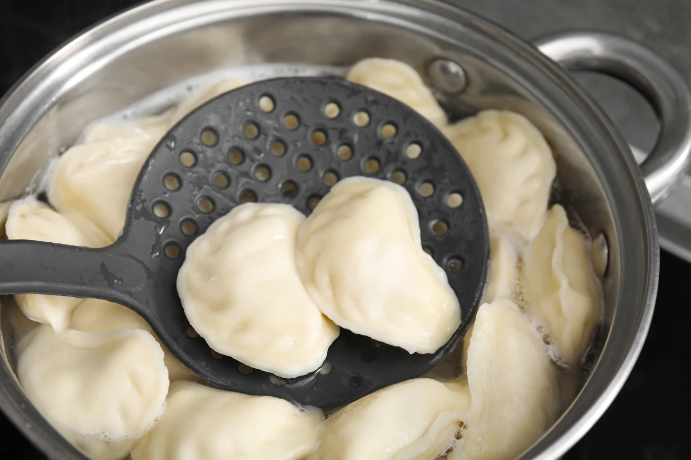 Pierogi w garnku podczas gotowania, podnoszone łyżką cedzakową. Dumplings unoszą się na powierzchni wody.