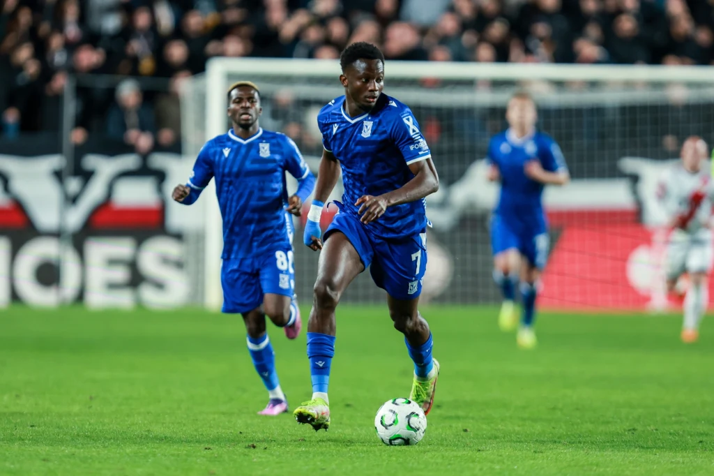 Yannick Agnero i Taofeek Ismaheel w barwach Lecha Poznań Piłkarz w niebieskim stroju prowadzi piłkę podczas meczu piłki nożnej, w tle widoczni inni zawodnicy i kibice na trybunach.