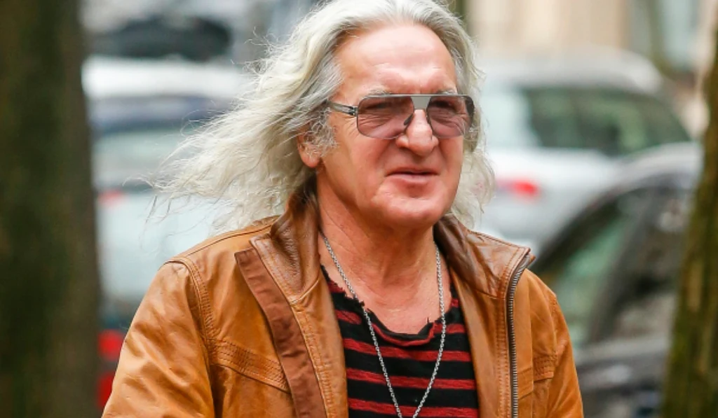 Starszy mężczyzna z długimi, siwymi włosami, w brązowej skórzanej kurtce i okularach przeciwsłonecznych, uśmiechnięty spaceruje po mieście. W tle widoczne zamazane samochody zaparkowane przy ulicy.