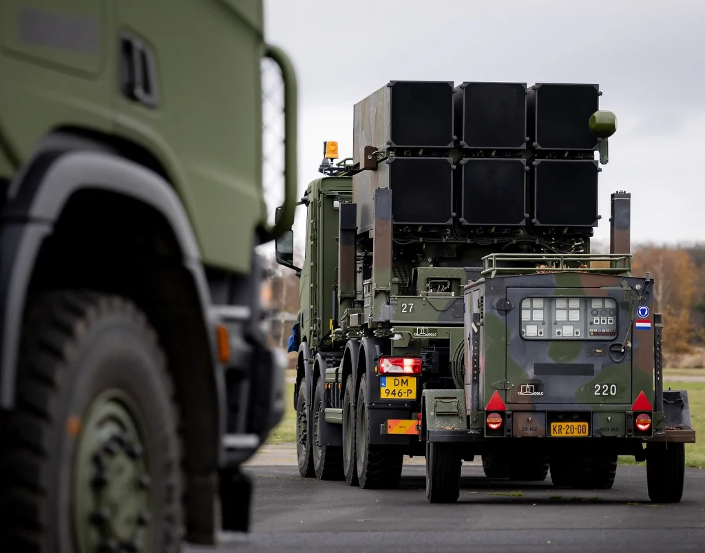 Wojskowy pojazd ciężarowy transportujący wyrzutnię rakietową ustawiony na asfaltowej drodze, z widocznymi szczegółami konstrukcji oraz dodatkowymi pojazdami wojskowymi w tle, w otoczeniu jesiennego krajobrazu.