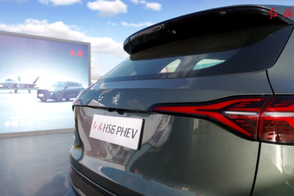 Tylny fragment zielonego samochodu Hongqi HS6 PHEV z widocznym oznaczeniem modelu i wyraźnym oświetleniem tylnym, w tle ekran z grafiką innych samochodów na tle nieba.