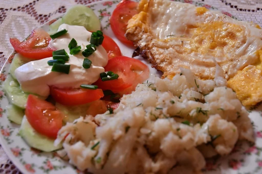 Porcja jajecznicy z dodatkiem gotowanych ziemniaków posypanych świeżym koperkiem oraz sałatka z ogórków i pomidorów z kleksem śmietany i szczypiorkiem na ozdobnym talerzu.