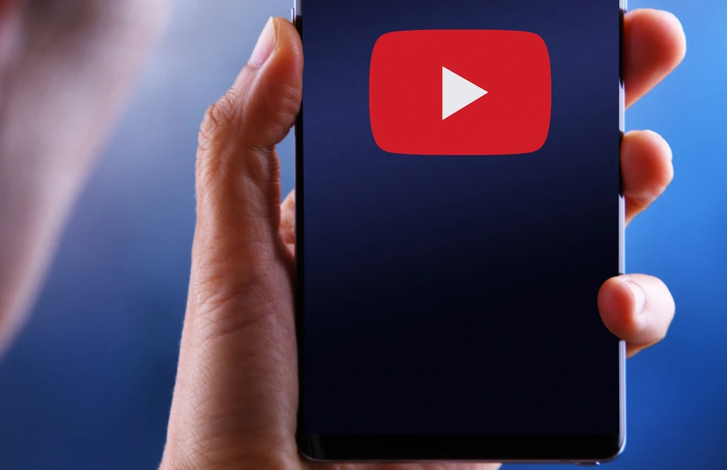 Dłoń trzyma smartfon z wyświetlonym dużym logotypem YouTube na ekranie, tło rozmyte w odcieniach niebieskiego.