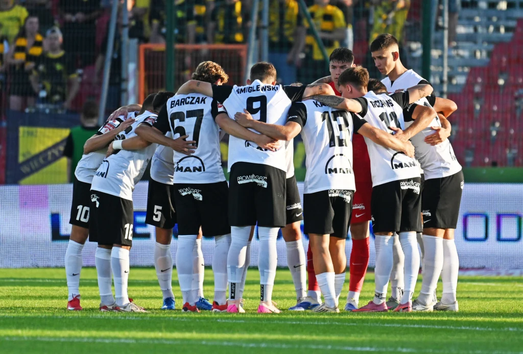 Grupa piłkarzy w strojach meczowych tworzy krąg, obejmując się wzajemnie na murawie stadionu. Zawodnicy przygotowują się do rozpoczęcia rozgrywki, a ich twarze skierowane są do środka. W tle widoczne są trybuny i rozmyta publiczność.