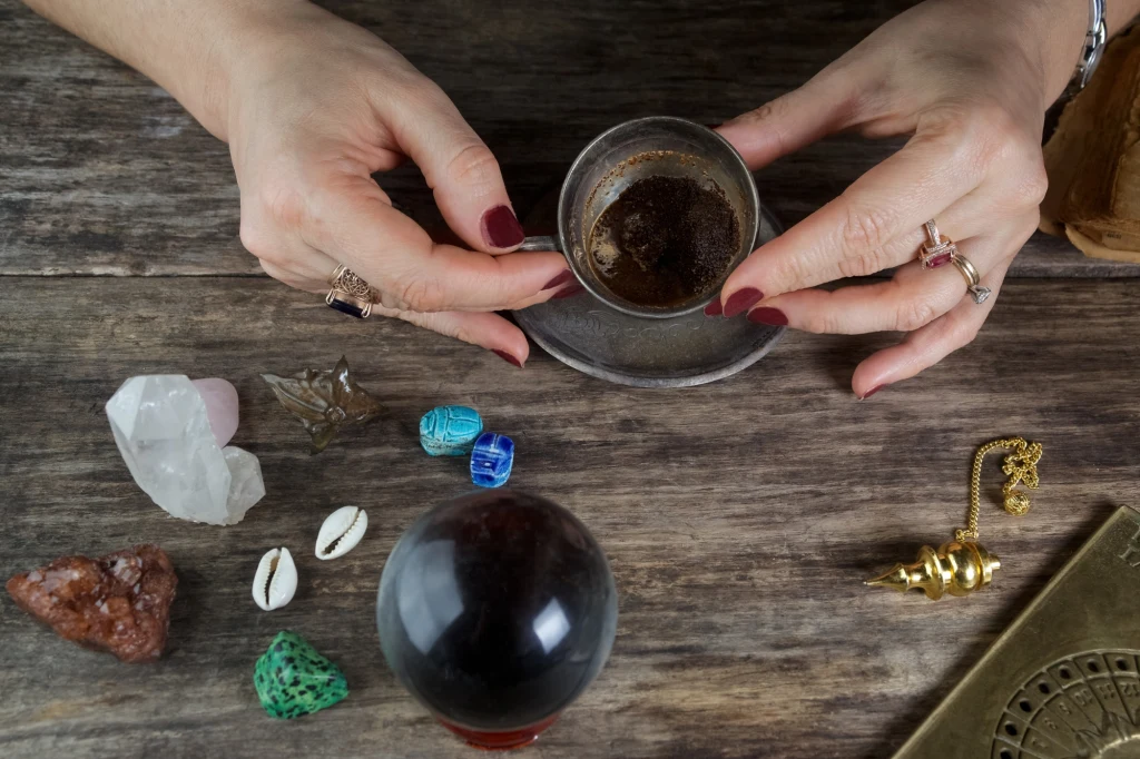 Ręce osoby trzymającej filiżankę z resztkami kawy otoczone różnorodnymi akcesoriami do wróżenia, w tym kryształami, muszlami, szklaną kulą, figurkami oraz wahadełkiem leżącymi na drewnianym stole.