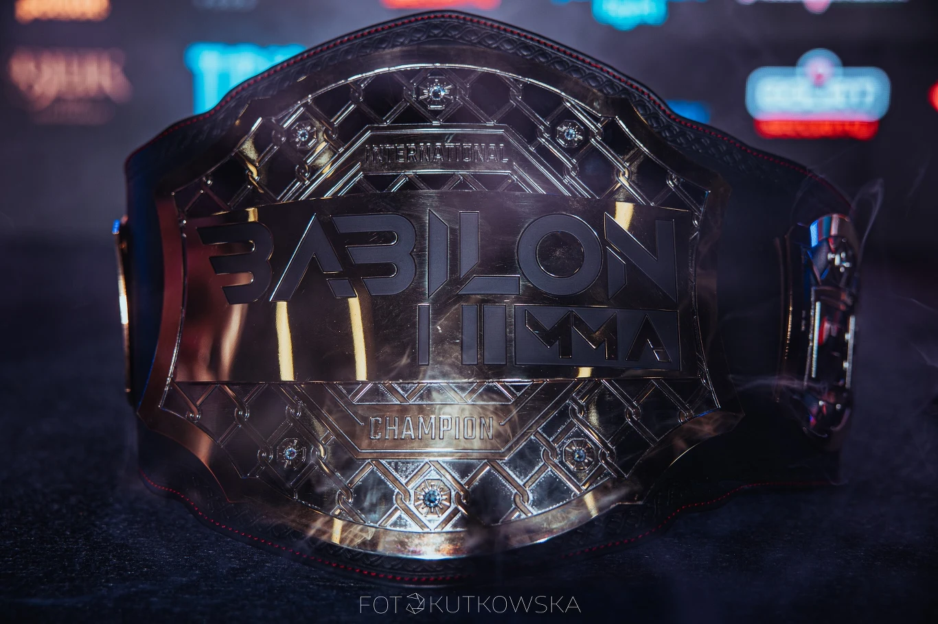 Pas mistrzowski Babilon MMA w wyraźnych złotych barwach, ustawiony na ciemnym tle z delikatnym odbiciem światła, na tle rozmazanych logotypów partnerów i organizatorów.