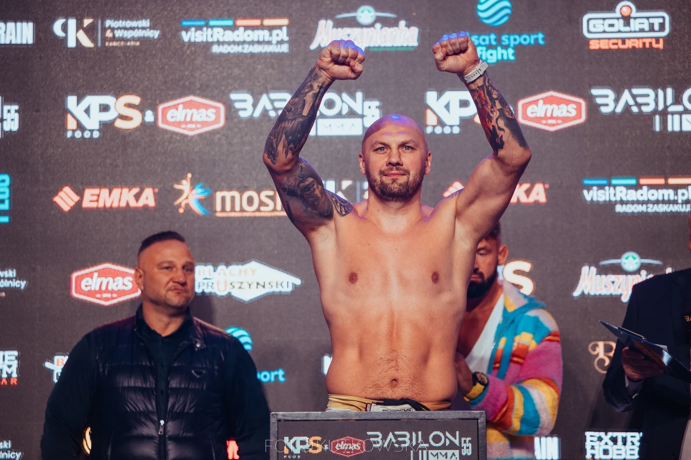 Umięśniony mężczyzna z tatuażami na ramionach stoi bez koszulki z uniesionymi rękami w geście triumfu, za nim widoczny jest tłum osób oraz tło ze sponsorami i logotypami gali sportowej.