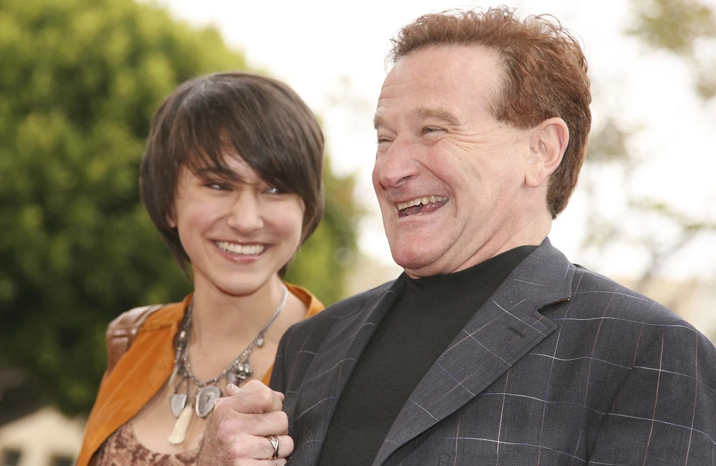 Dwie osoby, mężczyzna i młoda kobieta, śmieją się, stojąc blisko siebie na świeżym powietrzu, mężczyzna ubrany w marynarkę w kratę i czarny golf, kobieta w brązowej kurtce oraz z naszyjnikiem na szyi.