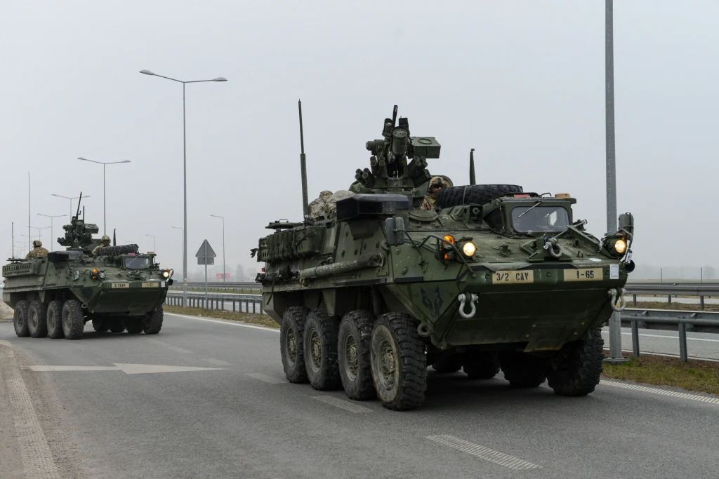 Dwa opancerzone pojazdy wojskowe poruszające się po asfalcie wzdłuż barier ochronnych, otoczone przez lampy uliczne oraz oznakowanie drogowe, widoczne w lekkiej mgle.