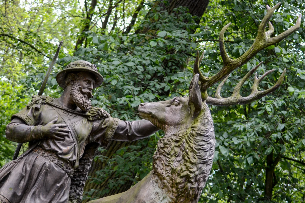Monument myśliwego w stroju z dawnych czasów z brodą i kapeluszem, stojącego obok jelenia o rozłożystych porożach, obaj otoczeni gęstą zielenią drzew i liści, wyrażają spokój i harmonię z naturą.