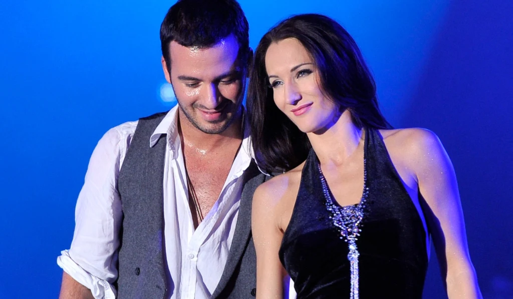 Para dorosłych osób ubrana elegancko, mężczyzna w białej koszuli i szarej kamizelce oraz kobieta w czarnej, błyszczącej sukience z efektownym naszyjnikiem, oboje uśmiechnięci na tle niebieskiego światła scenicznego.