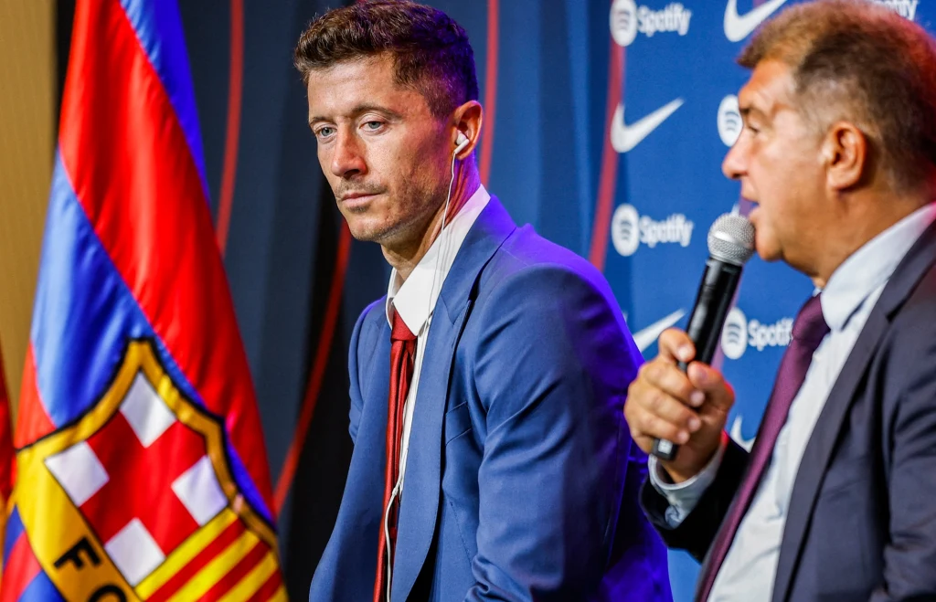 Robert Lewandowski i FC Barcelona szykują się do powrotu na Camp Nou Dwóch mężczyzn w garniturach siedzi obok siebie podczas konferencji prasowej, z widocznym mikrofonem w ręku jednego z nich, obok kolorowa flaga oraz logo klubu FC Barcelona.
