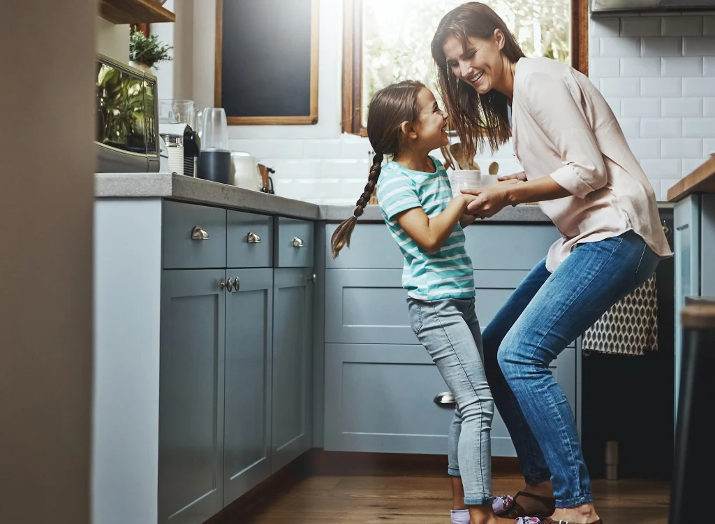 Matka w jasnej bluzce i dżinsach oraz córka w pasiastym t-shircie i sandałach, trzymają się za ręce i uśmiechają do siebie, stojąc w jasnej kuchni, światło słoneczne wpada przez okno, tworząc ciepłą, radosną atmosferę.