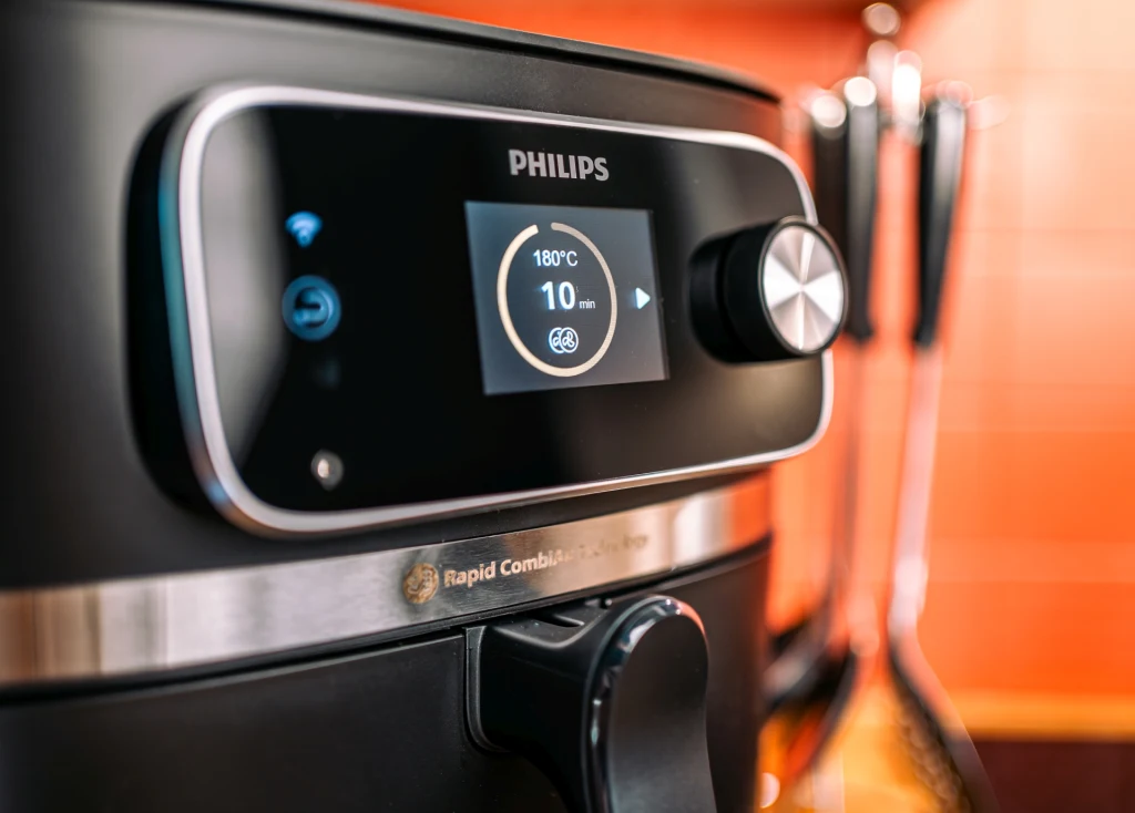 Nowoczesna frytkownica beztłuszczowa marki Philips z cyfrowym wyświetlaczem ustawionym na temperaturę 180°C i czas 10 minut, uchwyt do otwierania urządzenia oraz widoczne w tle przybory kuchenne.