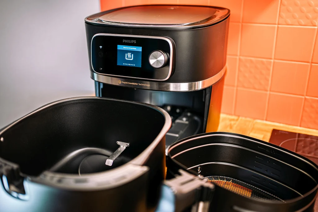 Czarna frytkownica beztłuszczowa marki Philips z otwartym koszem i panelem sterowania z wyświetlaczem cyfrowym, stojąca na blacie przy pomarańczowej ścianie pokrytej płytkami ceramicznymi.