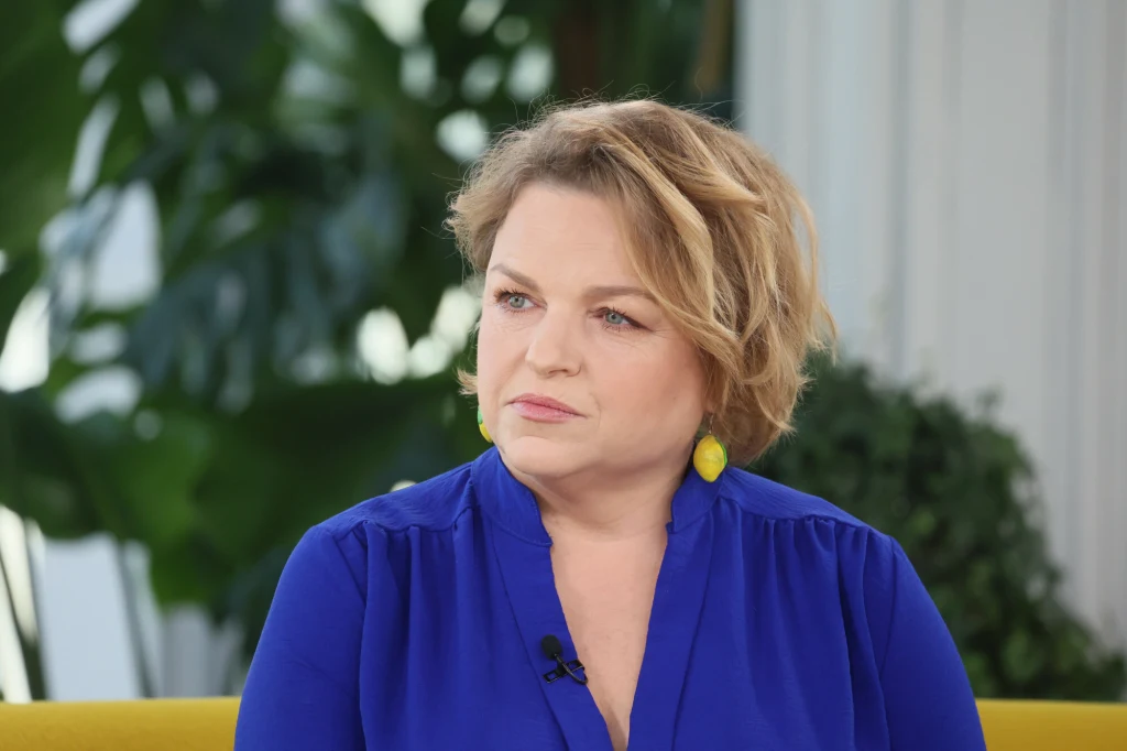 Katarzyna Bosacka wzięła pod lupę popularne ketchupy. Zamiast cukru zawierają one sztuczny słodzik Kobieta o blond włosach i krótkiej fryzurze ubrana w niebieską bluzkę siedzi na żółtej sofie. Na uszach ma żółte kolczyki, a za nią widać rośliny doniczkowe oraz białe jasne tło.