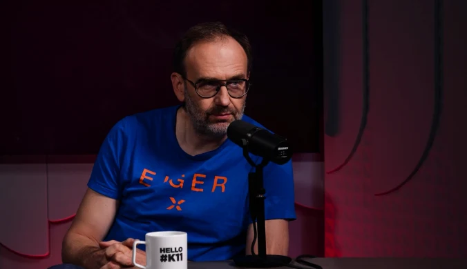 Mężczyzna w okularach siedzi przed mikrofonem w studiu, ubrany w niebieską koszulkę z napisem, trzyma kubek z napisem HELLO #KTI, tło ciemne z czerwonym podświetleniem.