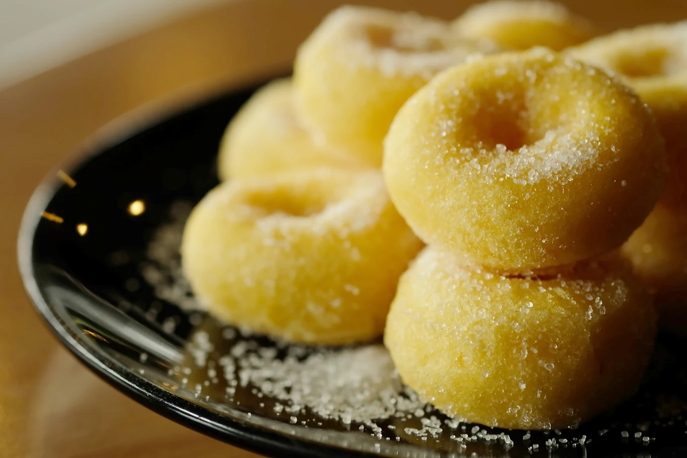 Stos małych, żółtych pączków posypanych cukrem ułożony na czarnym talerzu.