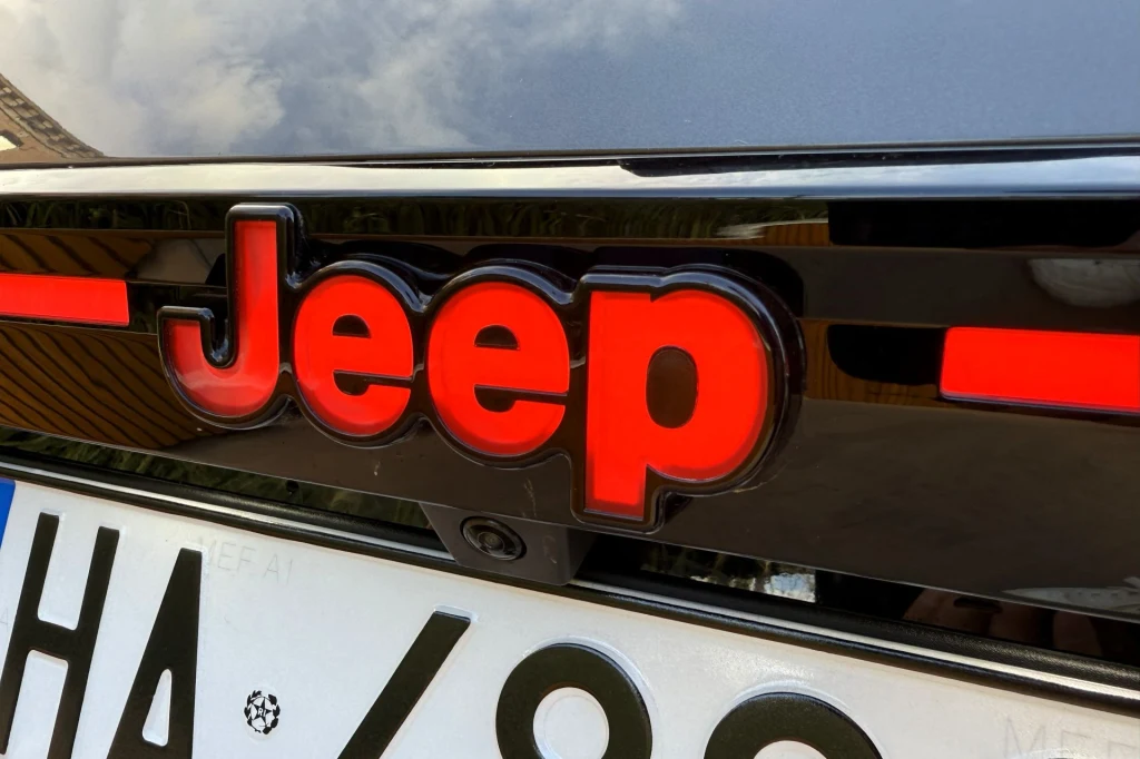 Na środku klapy bagażnika znajdziemy podświetlane logo Jeepa. Logo Jeep podświetlone czerwonym światłem na karoserii samochodu, pod nim znajduje się fragment tablicy rejestracyjnej.