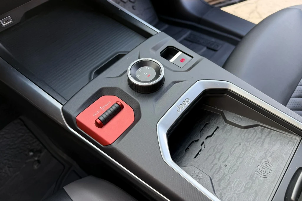 Masywne pokrętło skrzyni biegów i dźwignia zmiany trybów jazdy pasują do charakteru amerykańskiej marki. Panel centralny samochodu Jeep z widocznym pokrętłem sterującym, czerwonym przyciskiem trybu terenowego oraz podstawką na napoje i miejscem na drobne akcesoria.