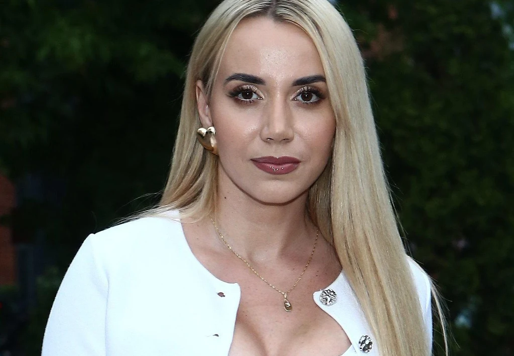 Młoda kobieta o długich blond włosach i wyrazistym makijażu, ubrana w elegancki biały strój z ozdobnymi guzikami oraz złotą biżuterią, stojąca na tle zieleni.