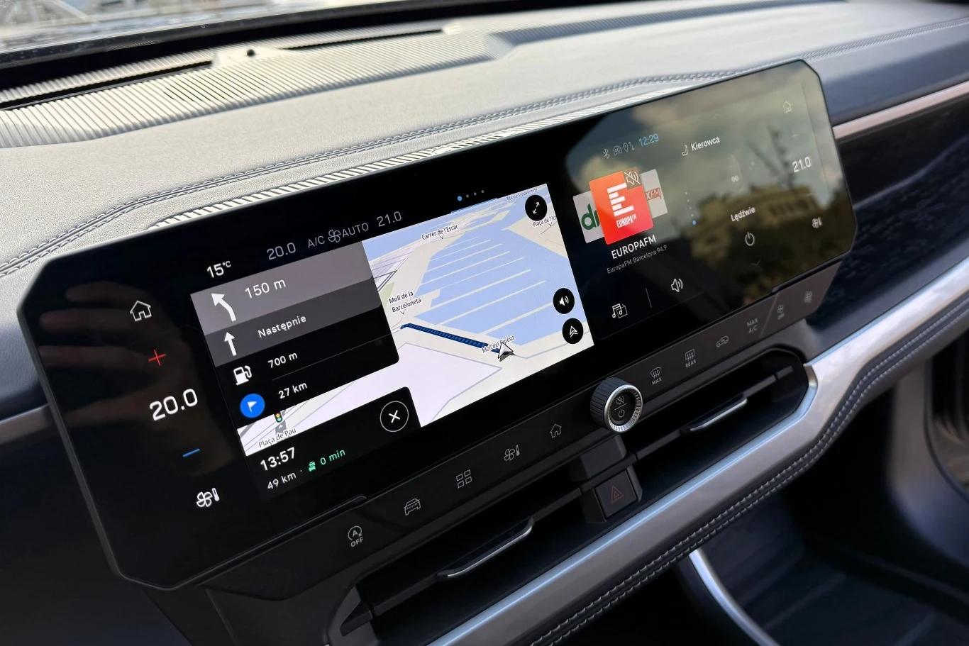 Kokpit samochodu z nowoczesnym panelem multimedialnym, wyświetlającym ekran nawigacji z trasą oraz dodatkowe informacje dotyczące ustawień pojazdu i pogody. Elegancki design oraz dotykowe przyciski pod ekranami podkreślają nowoczesność wnętrza.