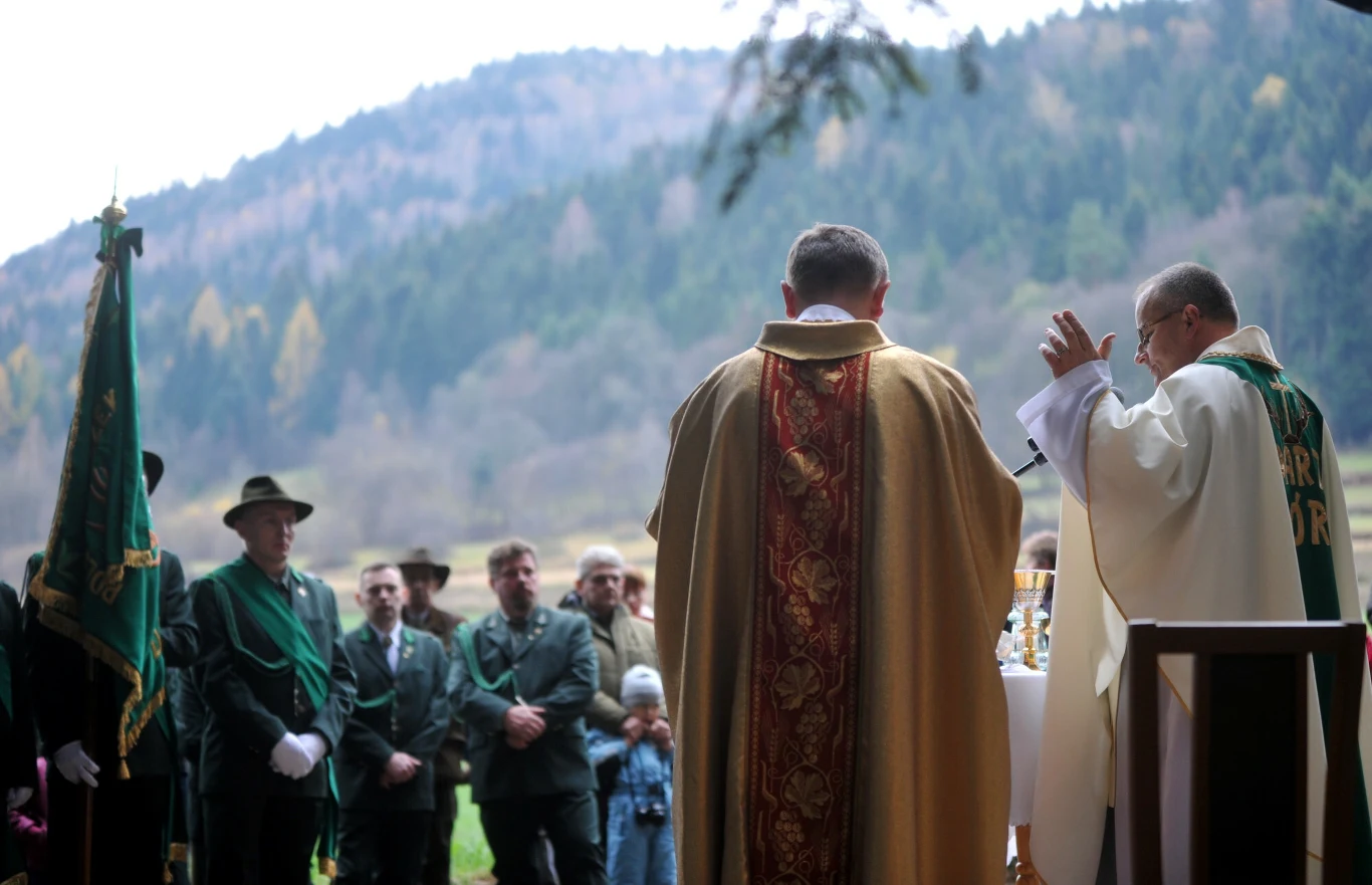 Grupa osób uczestniczy w mszy polowej prowadzonej przez dwóch księży w otoczeniu górskiego krajobrazu, po lewej stronie widoczni mężczyźni w mundurach z zielonym sztandarem myśliwskim.