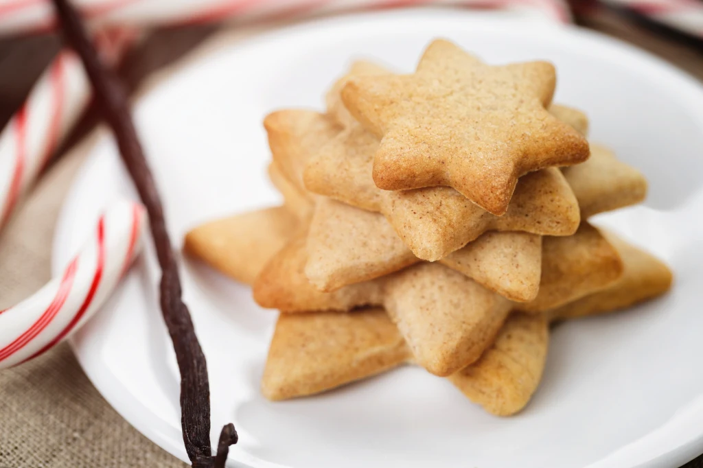 Stos gwiazdkowych ciasteczek ułożonych na białym talerzu, obok których znajdują się laski cukrowe i laska wanilii, wszystkie elementy utrzymane w świątecznym charakterze.