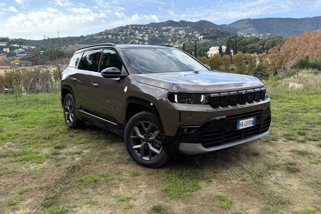 Jeep Compass jest dobrze wyciszonym autem. Podróżowanie nawet z prędkościami typowymi dla dróg szybkiego ruchu nie będzie obciążało naszego słuchu. Brązowy samochód terenowy marki Jeep stoi zaparkowany na trawiastym terenie, w tle znajdują się wzgórza porośnięte drzewami oraz zabudowania mieszkalne.