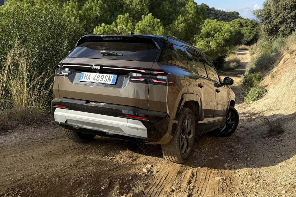 Nawet z napędem na przednią oś i trzycylindrowym silnikiem 1,2 Compass w terenie radzi sobie zaskakująco dobrze. Brązowy samochód terenowy marki Jeep porusza się po nierównej, piaszczystej drodze otoczonej gęstą zielenią drzew i krzewów, wyraźnie przystosowany do jazdy w terenie.