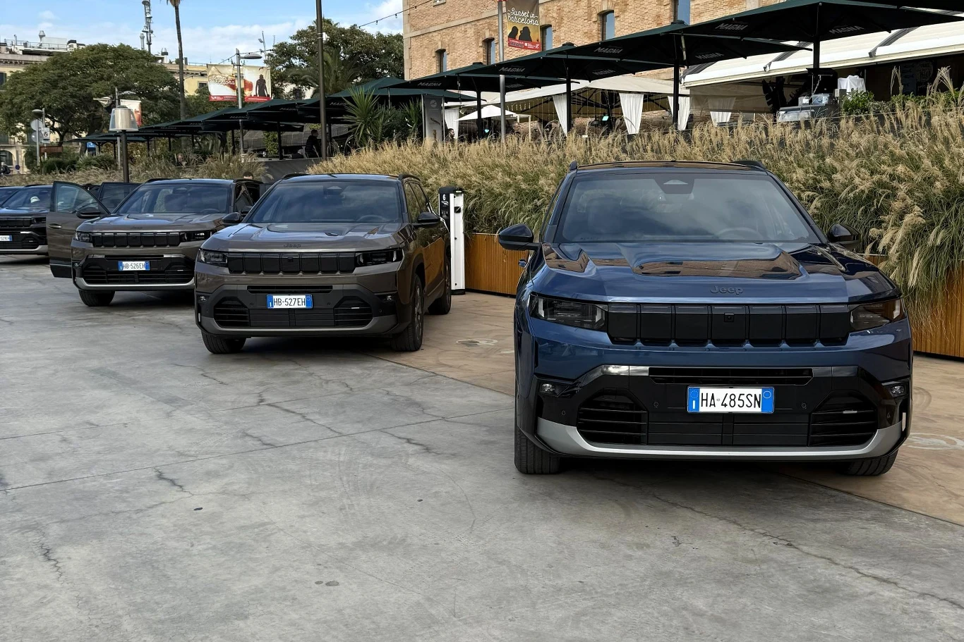 Trzy nowe SUV-y stojące w rzędzie na utwardzonym placu przy budynku, każdy pojazd ma włoską tablicę rejestracyjną, jasna pogoda i roślinność w tle.