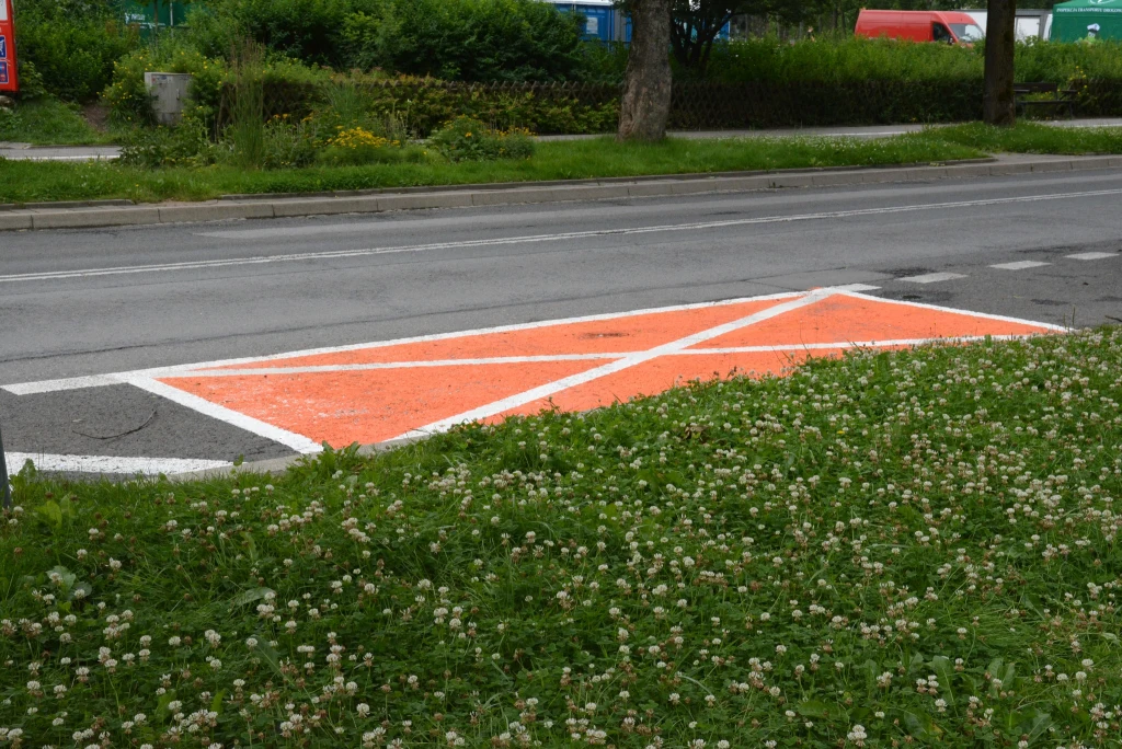 Pomarańczowa powierzchnia wyłączona z ruchu oznaczona białymi liniami na jezdni, otoczona zieloną trawą z kwiatami koniczyny, w tle asfaltowa droga i fragmenty zieleni miejskiej.