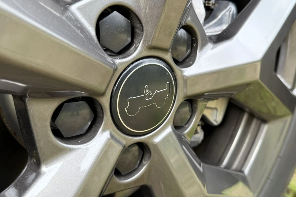 Jeep świetnie wykorzystuje swoje dziedzictwo. Przykładowo na dekielkach felg znajdziemy sylwetkę Willysa. Aluminiowa felga samochodowa z logo przedstawiającym sylwetkę klasycznego pojazdu terenowego. Widoczne szczegóły śrub montażowych oraz fragment tarczy hamulcowej.