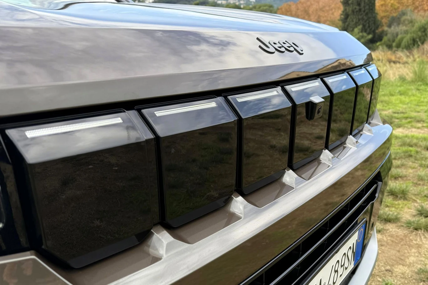 Zbliżenie na przedni grill samochodu marki Jeep z charakterystycznym logo, wyraźnym wzornictwem oraz fragmentem tablicy rejestracyjnej, na tle zielonego krajobrazu.