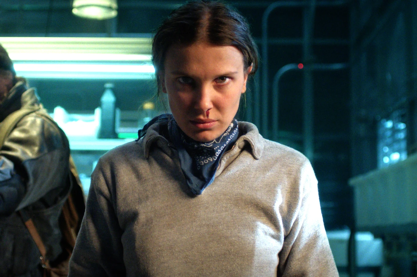 "Stranger Things" Jedenastka ze Stranger Things w szarej bluzie i chuście, w laboratorium, oświetlona niebieskawym światłem.