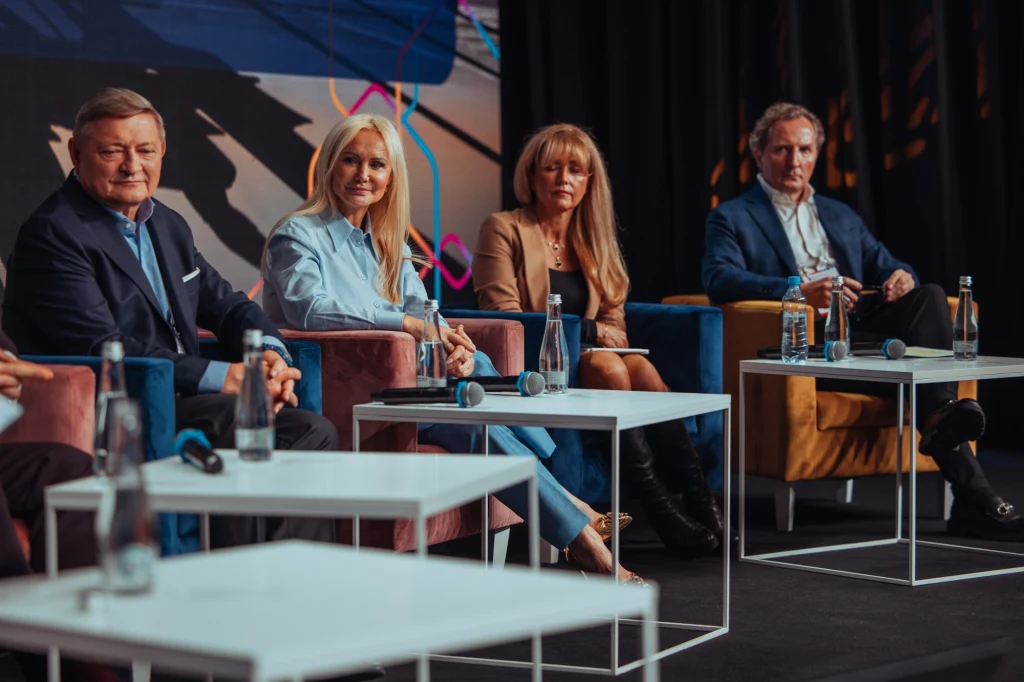 Czworo dorosłych, elegancko ubranych osób siedzi na scenie podczas panelu dyskusyjnego, przed nimi stoliki z wodą mineralną i mikrofonami, w tle kolorowa grafika.