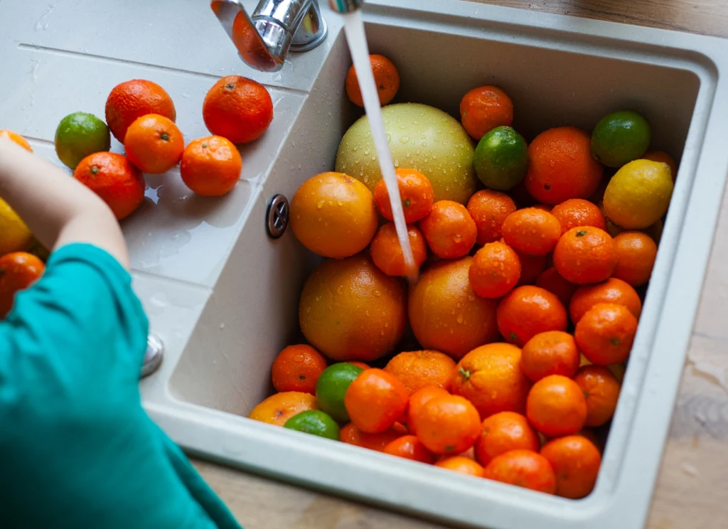 Duża ilość cytrusów, takich jak mandarynki, grejpfruty, pomarańcze i limonki, leży w zlewie, przy czym z kranu płynie strumień wody, a ręka osoby myje owoce.