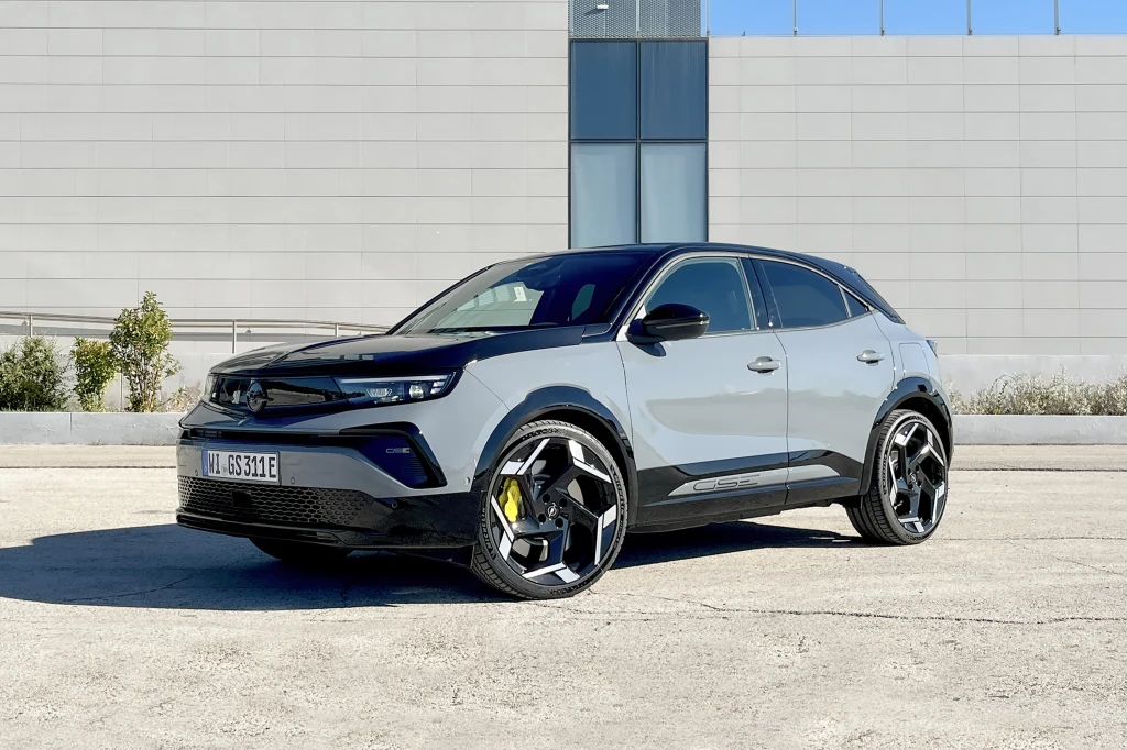 Nowoczesny szary samochód osobowy typu SUV stoi na pustym betonowym placu przed minimalistycznym budynkiem o jasnej elewacji. Auto wyróżnia się dużymi felgami, przyciemnianymi szybami oraz wyraźnie zaakcentowanymi liniami nadwozia.