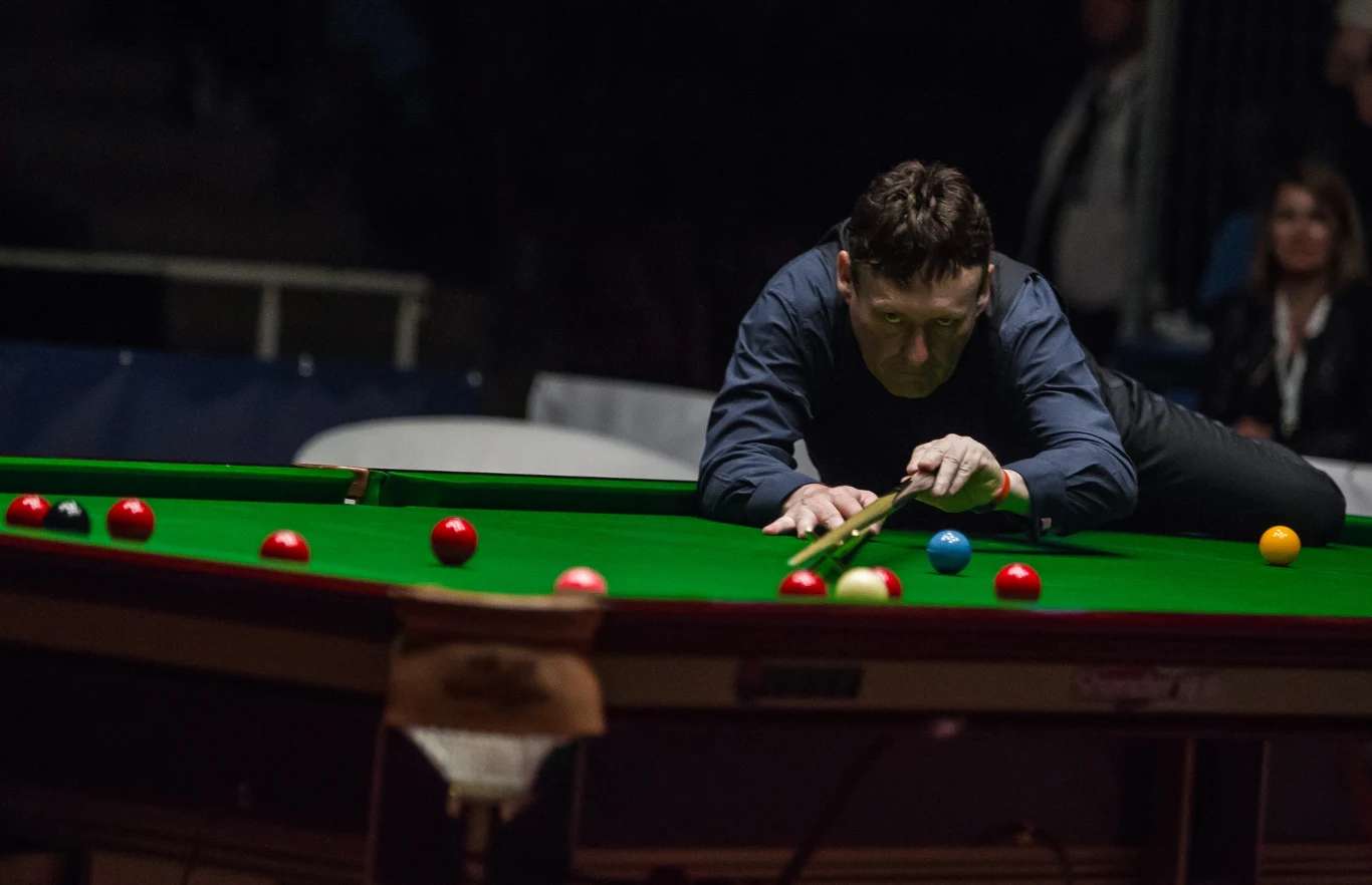 Zawodnik skupiony przy stole bilardowym w trakcie celowania kijem do snookera, wokół ciemne tło i widownia obserwująca grę.