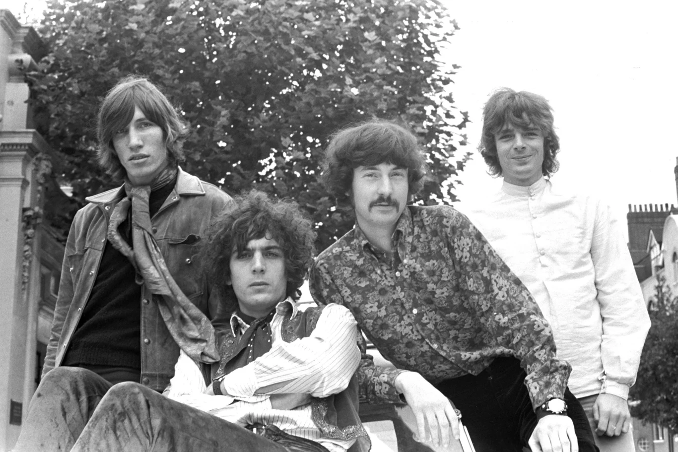 Pink Floyd w połowie lat 60. - od lewej: Roger Waters, Syd Barrett, Nick Mason i Richard Wright Czterech młodych mężczyzn stoi razem na tle drzew i nieba, ubrani w charakterystyczne dla lat 60. kolorowe i wzorzyste ubrania, prezentując spokojną i swobodną postawę.