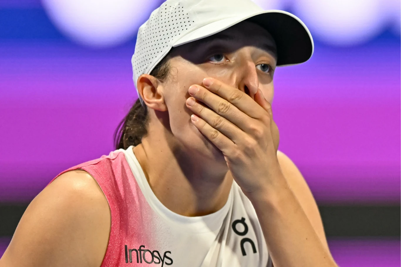 Młoda kobieta w sportowym stroju, z czapką z daszkiem, trzyma dłoń na ustach. Tło jest rozmyte w odcieniach fioletowego, a na jej twarzy widać emocje, prawdopodobnie zaskoczenie lub stres.