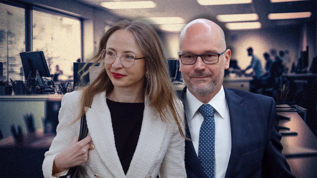 Dwie osoby w eleganckich ubraniach biznesowych stoją razem w przestrzeni biurowej, w tle widoczne komputery i biurka oraz sylwetki pracowników w biurze open space.