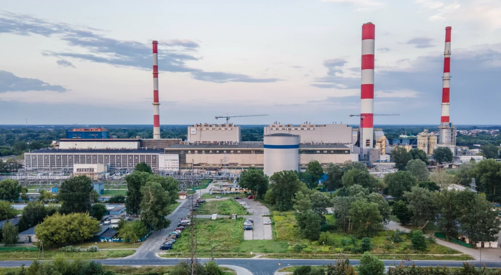 Elektrownia z trzema wysokimi kominami w biało-czerwone pasy, otoczona zielenią oraz niską zabudową miejską. Na pierwszym planie widoczne są drogi, drzewa i kilka budynków mieszkalnych