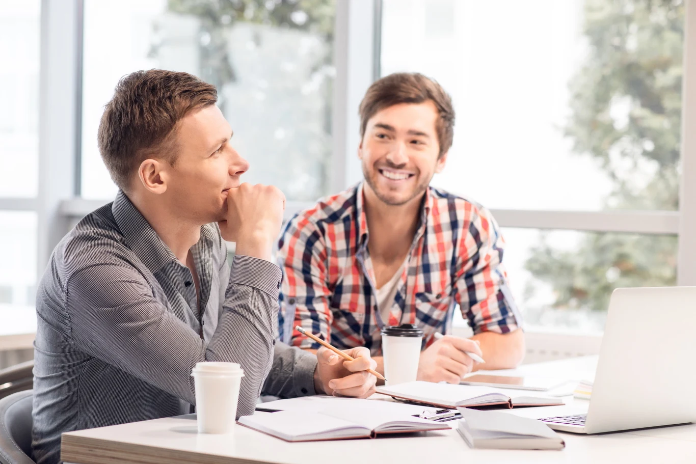 O zdrowie warto pytać. Niby dla kolegi, a tak naprawdę dla siebie Dwóch młodych mężczyzn rozmawiających przy stole w nowoczesnym biurze, z kubkami kawy, notatkami i laptopem przed sobą, jasne tło z dużymi oknami.