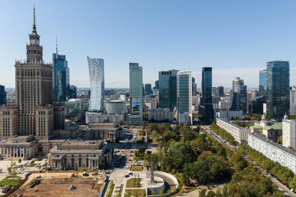 Panorama centrum Warszawy z widokiem na Pałac Kultury i Nauki oraz nowoczesne wieżowce i otaczającą zieleń miejską, w tle błękitne niebo.