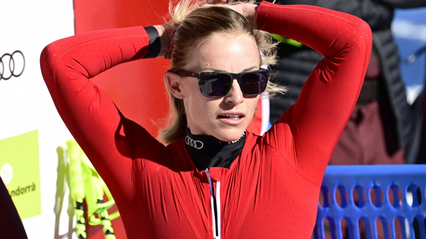 Lara Gut-Behrami Kobieta w sportowym czerwonym kombinezonie i okularach przeciwsłonecznych poprawia włosy, stojąc na tle barierki i tablicy z logotypami.